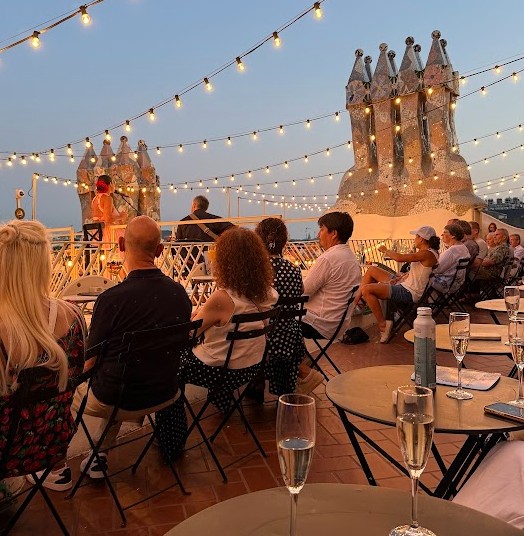 Due file di tavolini e gente seduta con il proprio calice di cava che ascolta il concerto chitarra e voce sul tetto di Casa Batllò per le magic nights