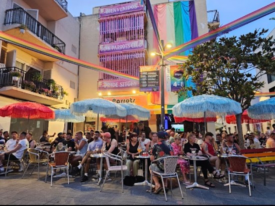 Tavolini all'aperto del Parrots Pub Sitges, in una piazzetta decorata con striscioni arcobaleno